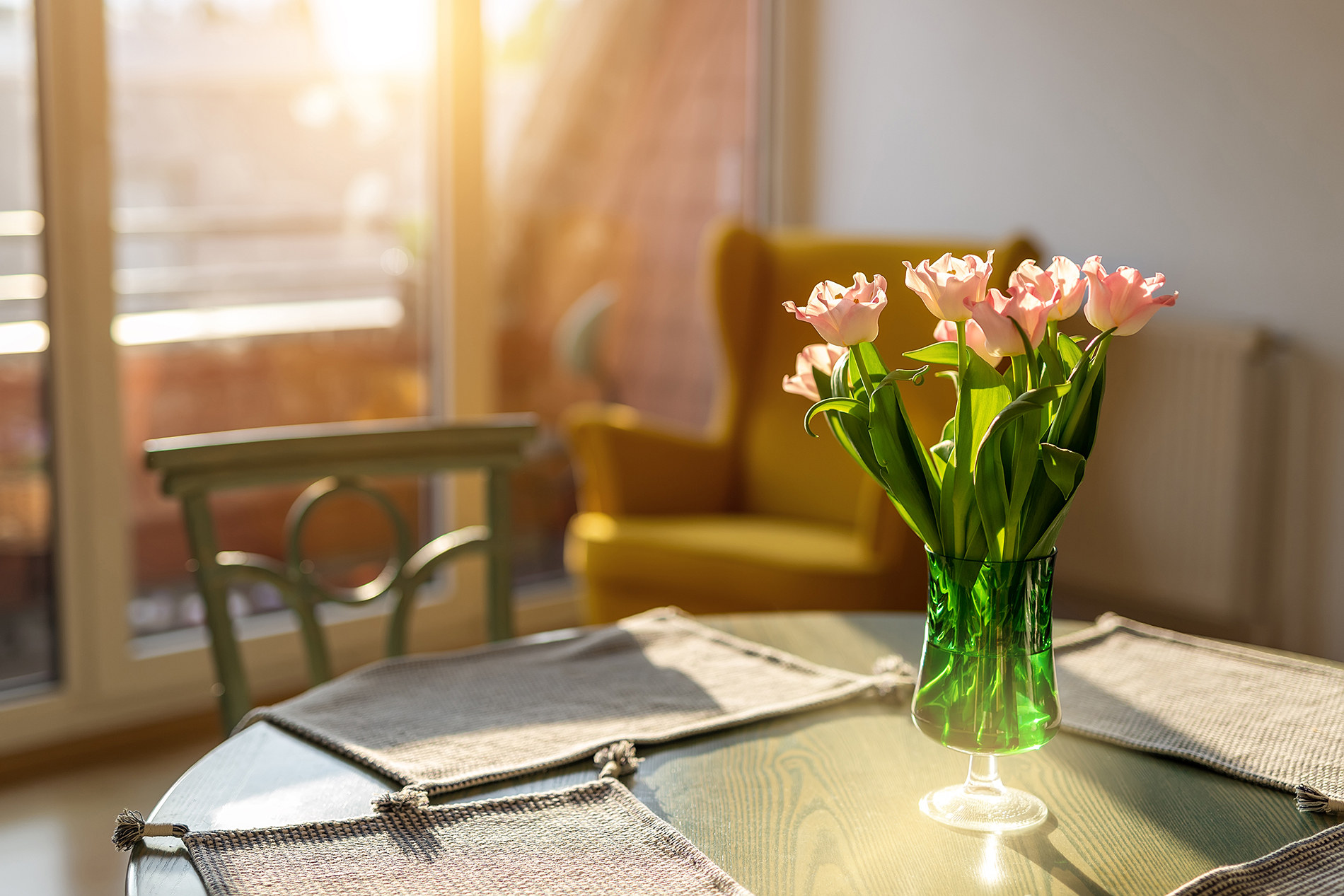 Beautiful fresh pink tulips bouquet in green glass vase on table in warm sunset sun lights against balcony window in cozy home interior. Blooming flowers decoration in living room provence style.